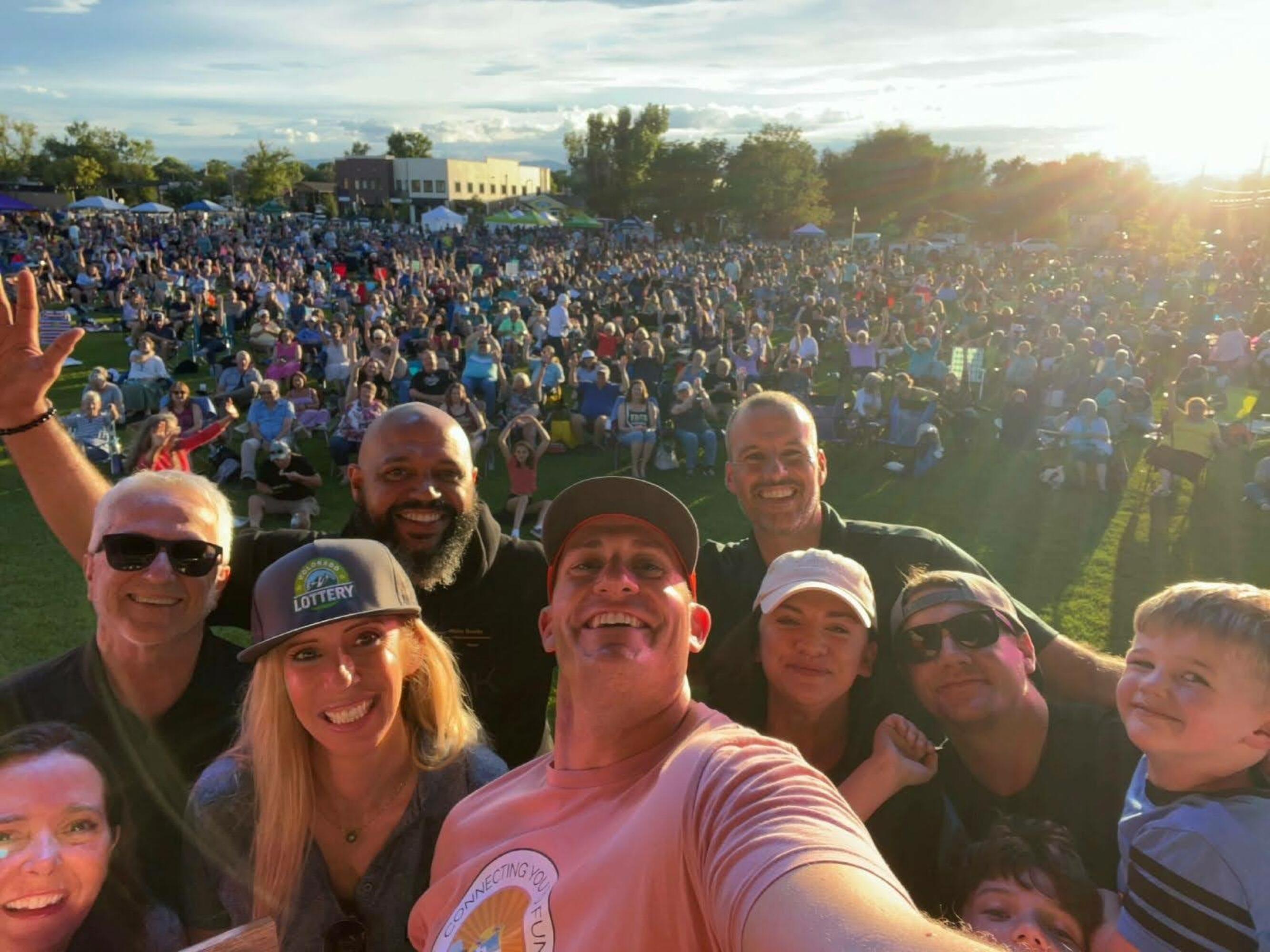 A group of smiling people take a selfie in the foreground while a large crowd of concertgoers sits on a grassy field behind them during a sunny outdoor event.